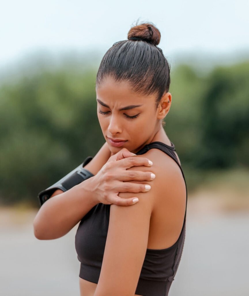 Young woman pausing during a workout, experiencing shoulder pain, which may indicate inflammation. She is outdoors, looking down at her shoulder with a concerned expression, highlighting the symptoms of pain and discomfort associated with inflammation.