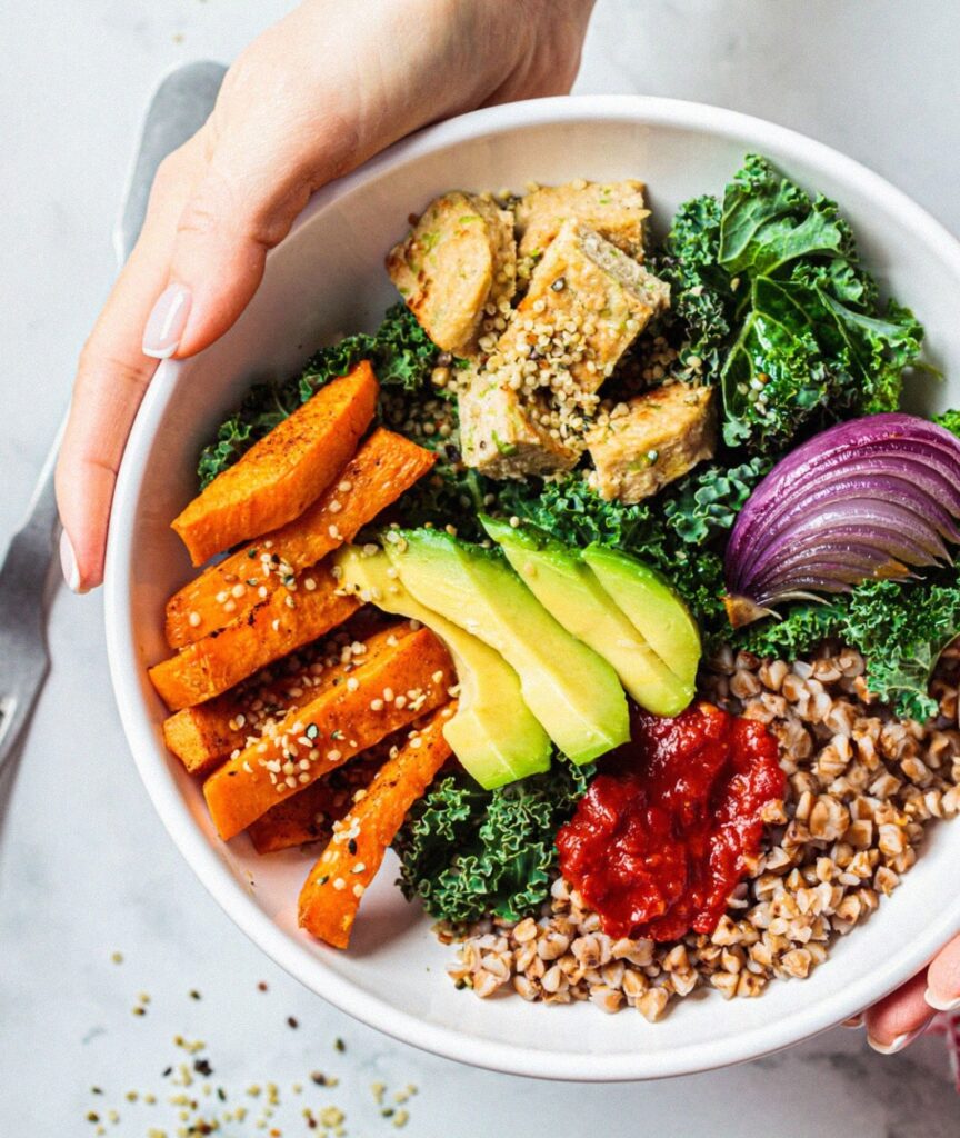 Colorful plant-based meal in a bowl, showcasing a healthy array of ingredients including roasted sweet potato wedges, sliced avocado, grilled tofu, kale, cooked buckwheat, and a dollop of red sauce. A person's hand is seen holding the bowl, emphasizing a conscious choice towards healthier, inflammation-reducing food options.