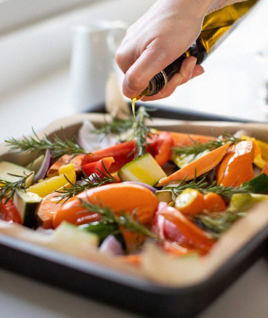 Close-up of a person drizzling olive oil over a tray of colorful chopped vegetables including bell peppers, zucchini, and red onions, garnished with fresh rosemary. This image highlights the use of high-quality olive oil as a healthy, heat-stable fat source for cooking to reduce inflammation.