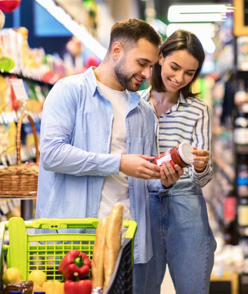 A young couple smiling while examining a jar of sauce in a grocery store aisle, illustrating a mindful approach to shopping with a focus on reducing sugar intake. The woman is pointing at the nutritional label, emphasizing the importance of checking food labels for added sugars to maintain a healthy diet.