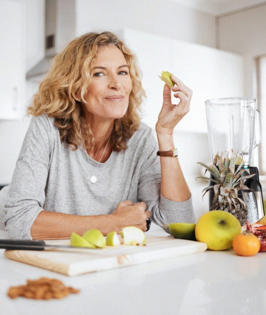 A mature woman with curly hair smiling at the camera, holding a slice of green apple in her kitchen. She is surrounded by various fruits and a blender, illustrating a lifestyle focused on personalized nutrition and testing different foods to manage inflammation.