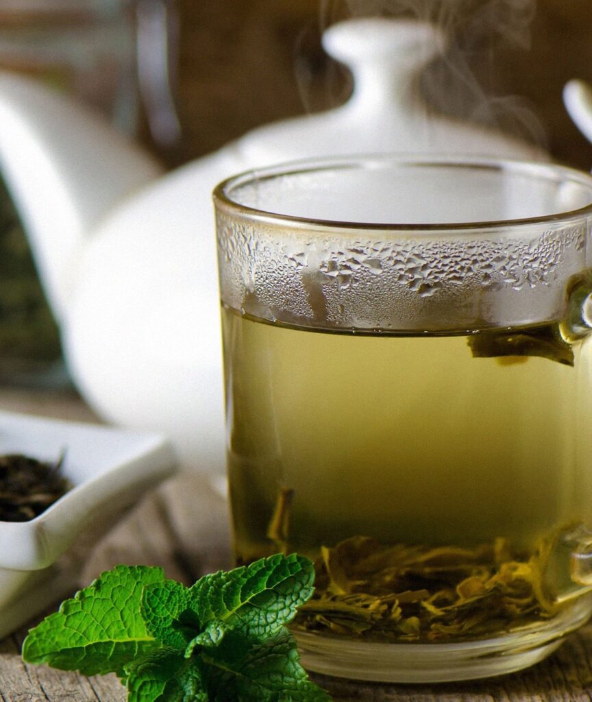 Close-up of a steaming cup of green tea accompanied by fresh mint leaves, set against a rustic wooden table with a tea pot in the background. This image illustrates a soothing, health-promoting beverage known for its anti-inflammatory properties due to its rich antioxidant content.